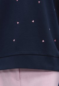 Υφασμα σε ναυτικό μπλε με μικρά ροζ μοτίβα καρδιών πάνω από ένα ριμπ ύφασμα στο τελείωμα και ένα ανοιχτό ροζ κάτω ένδυμα ορατό από κάτω.