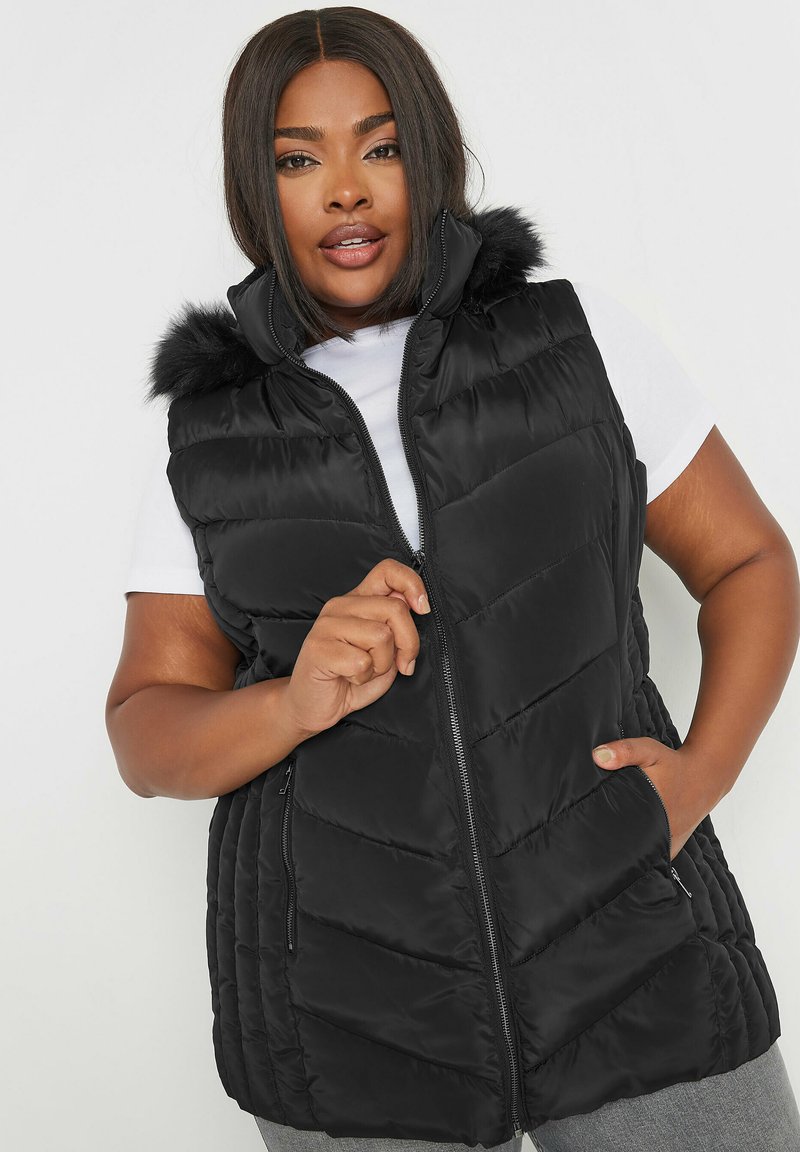 Mustapuffertakki, jossa on tekoturkistettu huppu, neljäkulmainen muotoilu, vetoketjusuljenta ja sivutaskut. Päällä valkoinen yläosa.