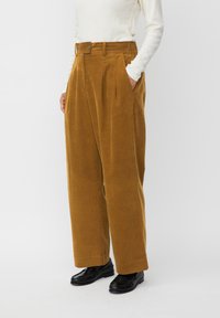 Pantaloni in corduroy color carne con vita alta, gamba larga e pieghe. Presenta tasche laterali e chiusura a pulsante. Indossati con scarpe nere.