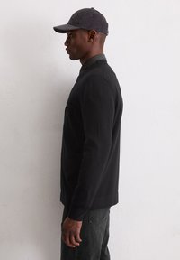 Schwarzer Pullover mit rundem Ausschnitt und gerippten Bündchen, darüber ein kariertes Hemd getragen. Ein graues Cord-Cap rundet den Look ab.