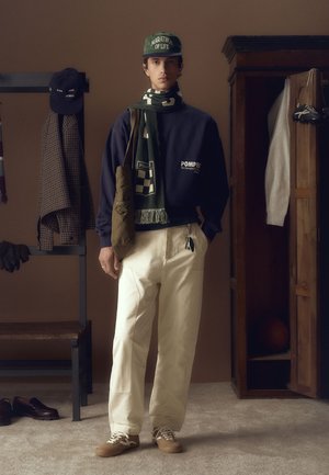 Marineblauer Sweatshirt mit Logo, grüner Schal mit Muster, cremefarbene Hose, beigefarbene Schuhe. Accessoires umfassen eine Mütze und eine Umhängetasche. Neutraler Hintergrund.