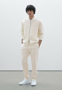 Veste zippée en crème clair avec des poignets côtelés, assortie à un pantalon fuselé. Des baskets blanches complètent cette tenue minimaliste. Texture de tissu lisse.