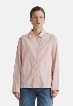 Mujer con una camisa de manga larga de color rosa claro con cuello y vaqueros azules, de pie frente a un fondo blanco liso.