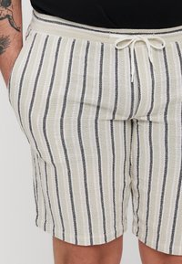 Shorts beiges avec des rayures verticales noires et blanches, dotés d'une taille élastique avec cordon de serrage et de poches latérales. Tissu texturé.