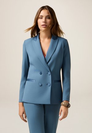 Blazer bleu à double boutonnage avec col cranté, pantalon ajusté assorti, texture lisse et boutons décoratifs, porté avec un haut rouge en dessous.