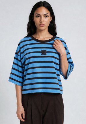 Young woman with long dark hair wears a loose blue and black striped shirt with a small decorative emblem on the chest.
