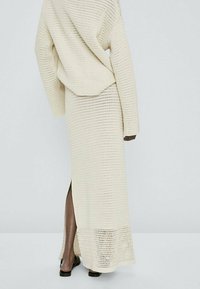 Personne portant une longue jupe en crochet couleur crème avec une fente au dos et un pull ample assorti, debout devant un fond clair uni.