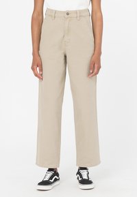 Pantaloni beige a vita alta con gamba larga, dotati di chiusura frontale a bottone e cerniera, realizzati in twill di cotone, abbinati a sneakers nere e bianche.