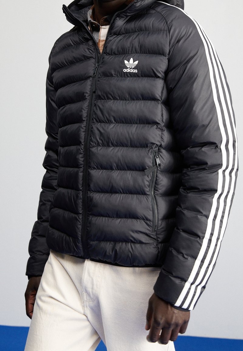 Černá péřová bunda s předním zipem a kapucí. Obsahuje bílé třípruhové akcenty na rukávech a logo Adidas na levé horní straně.