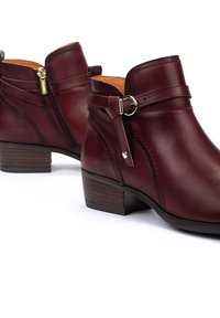 Botas de cuero burdeos hasta el tobillo con una cremallera lateral, correa decorativa con hebilla y un tacón de madera bajo, que presentan una textura suave y detalles de costura.