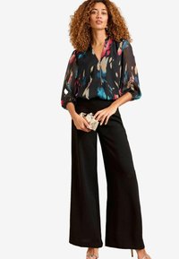 Pantaloni neri a gamba larga abbinati a una blusa nera trasparente con motivi astratti colorati e zip frontale. Accessoriati con una pochette geometrica.