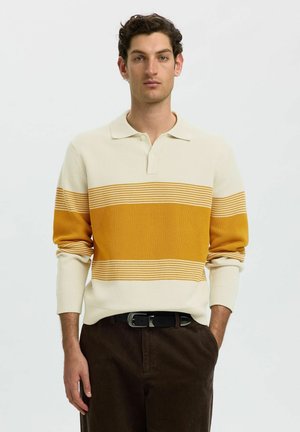 Jeune homme aux cheveux bouclés foncés portant un pull crème à manches longues avec des rayures jaunes et un pantalon marron foncé, debout avec une main dans la poche.