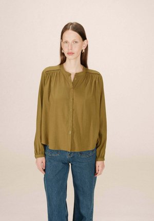 Chemisier vert olive à manches longues et encolure ronde, avec des fronces aux épaules. Associé à un jean bleu taille haute.