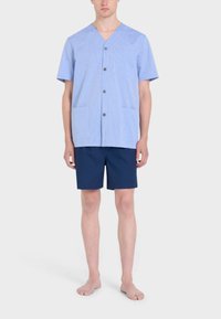 Camicia a maniche corte a righe blu chiaro con scollatura a V, due tasche frontali e bottoni, abbinata a pantaloni corti blu scuro. Modello a piedi nudi.