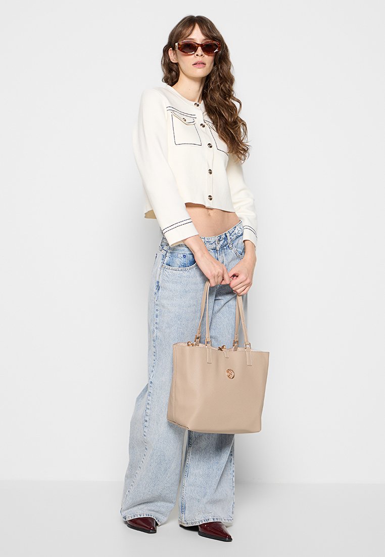 Sac à dos beige avec des anses robustes, texture en cuir lisse et détails piqués ; porté avec un jean large en denim clair et un top blanc court.