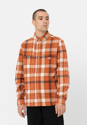 Chemise en flanelle à carreaux orange et blancs avec des accents marron, deux poches poitrine et des boutons-pression. Le modèle la porte avec un pantalon noir.