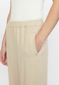 Beige weite Hosen mit elastischem Bund und Seitentasche. Hergestellt aus strukturiertem Stoff, mit einem dezenten Stickdetail in der Nähe der Tasche.