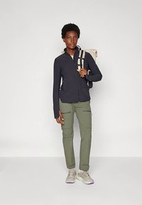 Vrouw in een zwarte ritsjack, olijfgroene cargobroek, lichtgekleurde sneakers en met een beige rugzak op één schouder.