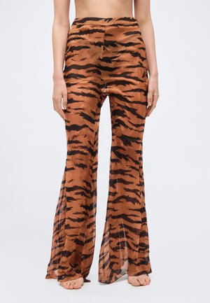 Femeie purtând pantaloni largi cu talie înaltă, transparenți, cu dungi de tigru, stând desculță cu mâinile relaxate pe lângă corp, pe un fundal simplu.