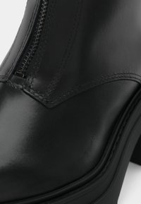 Close-up of a black leather boot featuring detailed stitching and a side zipper on the upper part of the shoe.