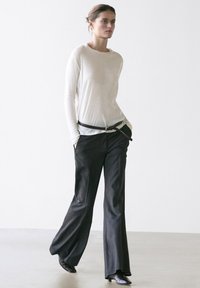 Kvinde går med en hvid langærmet top stoppet ned i sorte vidde bukser, iført et tyndt sort bælte og sorte spidse sko.