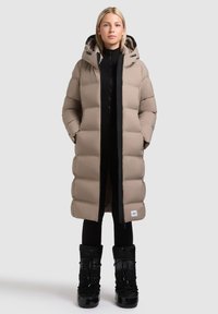 Beige, bauschiger, knielanger Mantel mit Kapuze, ausgestattet mit einem schwarzen Reißverschluss und seitlichen Taschen, kombiniert mit schwarzen isolierten Stiefeln.