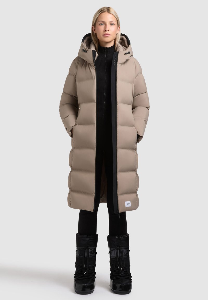 Beige, bauschiger, knielanger Mantel mit Kapuze, ausgestattet mit einem schwarzen Reißverschluss und seitlichen Taschen, kombiniert mit schwarzen isolierten Stiefeln.