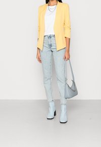 Blazer jaune, t-shirt blanc, jean taille haute bleu clair et bottines à talons bleu clair. Sac gris avec détail en chaîne. Textures lisses.