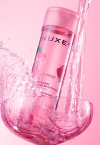 Transparent rosa micellärvattenflaska med ett rosa lock, märkt "NUXE Paris," som plaskar ner i en klar skål fylld med vatten.