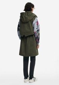 Une veste utilitaire vert olive avec un ourlet pointu, portée par-dessus un sweat-shirt gris avec des graphismes de dessins animés colorés et un jean noir.