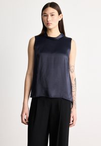 Tmavomodrý tielkový top z hladkej tkaniny s jemným leskom, s vysokým výstrihom a voľným strihom, nosený cez čierne nohavice s širokými nohavicami.