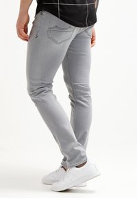 Jeans slim fit de color gris claro con bolsillos traseros, que presentan una textura suave y detalles de costura sutiles, combinados con zapatillas blancas.