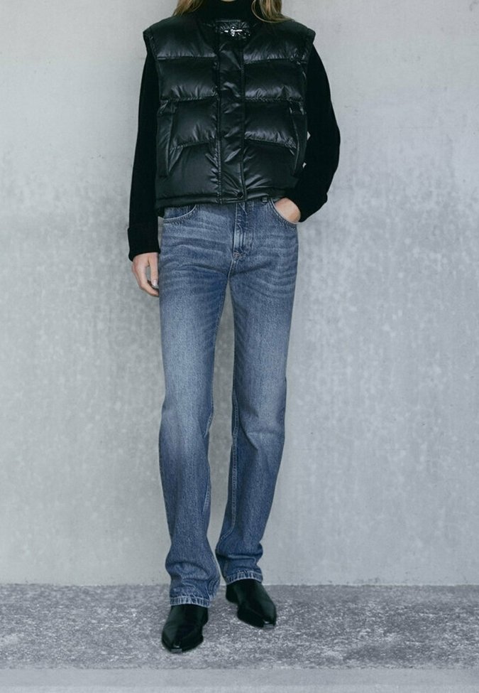 Personne portant une veste matelassée noire par-dessus un top noir à manches longues, un jean droit bleu et des chaussures pointues noires, debout contre un mur gris.