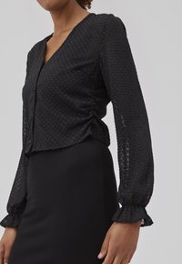 Blouse noire texturée avec un col en V, des manches longues bouffantes avec des poignets volants et un boutonnage à l'avant. Présente des détails froncés sur le côté.