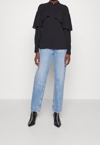 Blusa nera a rouches con colletto alto, abbinata a jeans dritti azzurri chiari e stivaletti neri a punta. Tessuto dalla consistenza liscia.