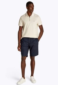 Polo crema a maniche corte con colletto a mezza cerniera, abbinata a pantaloncini blu navy e sneakers bianche. Tessuto liscio, stile classico, senza motivi visibili.