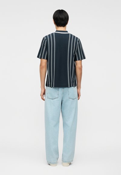 Άτομο που στέκεται πλάτη, φορώντας μαύρο κοντομάνικο πουκάμισο με κάθετες λευκές ρίγες και ανοιχτόχρωμο τζιν παντελόνι.