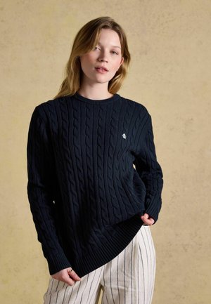 Giovane donna con capelli chiari indossa un maglione blu navy lavorato a trecce e pantaloni bianchi con sottili righe nere verticali su sfondo beige.
