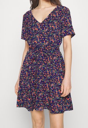 Žena má na sobě šaty s krátkým rukávem a výstřihem do V, sahající do kolen, s tmavě modrým pozadím a drobným květinovým vzorem v červené, zelené a fialové barvě.