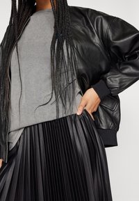 Svart läder bomberjacka med ribbade manschetter, grå sweatshirt under, och en svart plisserad kjol. Texturkontraster i tyger.