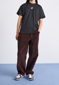 Persona vestida con una camiseta de malla negra de Nike, pantalones de pana marrones holgados y zapatillas blancas, negras y rojas, de pie sobre un suelo azul.