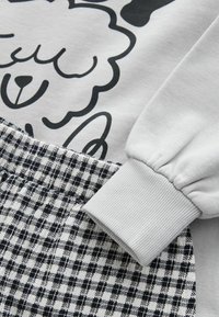 Sweat-shirt gris avec un graphisme noir de mouton en dessin animé, accompagné d'un pantalon à carreaux noir et blanc. Les poignets sont en côte et élastiqués.