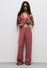 Pletený dvoudílný outfit v rezavé barvě, který zahrnuje halter crop top a kalhoty s širokými nohavicemi. Texturovaná látka s uvolněným střihem. Černé sluneční brýle a doplňky.