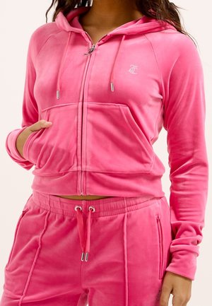Femme portant un sweat à capuche zippé en velours rose vif avec cordons et un pantalon assorti à cordons, les mains dans les poches.