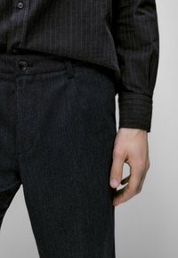 Dunkle Denimhosen mit glatter Textur, ausgestattet mit einem Knopfverschluss und Seitentaschen, kombiniert mit einem schwarz gestreiften Hemd.