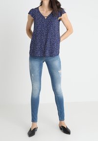 Donna che indossa una blusa corta a maniche corte floreale blu, jeans skinny leggermente strappati, e mule nere a punta, in piedi con le mani dietro la schiena.