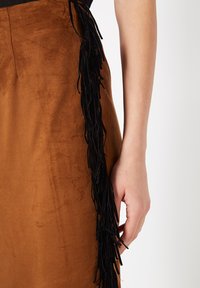 Falda de ante marrón con acabado texturizado, con un detalle de flecos negros a lo largo del lateral y cintura ajustada.