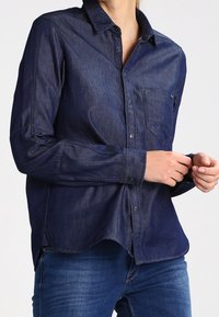 Mörkblå denimskjorta med lång ärm, knäppning fram, en enda bröstficka och något rundad nederkant. Slät textur med skräddarsydd passform.