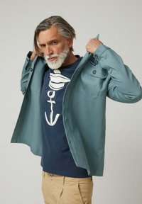 Camisa de color verde azulado claro abotonada con dos bolsillos delanteros, usada abierta sobre una camiseta azul marino con un diseño gráfico blanco. Pantalones beige completan el conjunto.