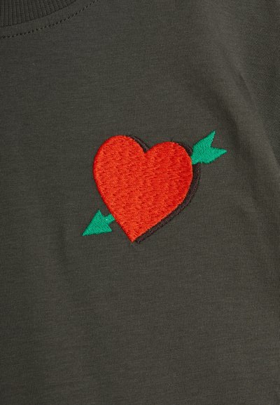 Olive green fabric featuring an embroidered red heart with a black outline, pierced by a green arrow, highlighting texture and contrast.